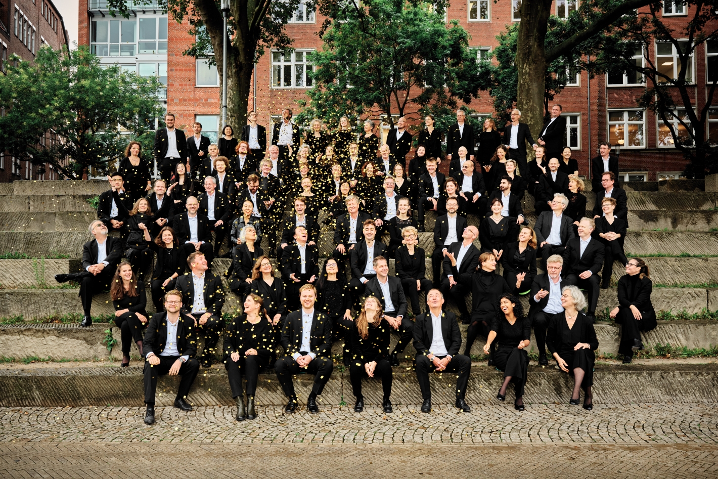 Die Bremer Philharmoniker auf einer Treppe sitzend, Goldregen fällt auf sie herab, Fotocredit Caspar Sessler